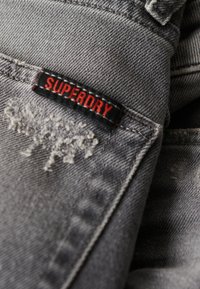 TelA de mezclilla gris con acentos desgastados. Presenta una etiqueta negra con "SUPERDRY" en rojo. Primer plano de los detalles de costura y textura.