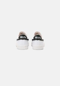Hvite sneakers med glatt stoffoverdel, svarte detaljer på hælen og strukturert gummisåle. Enkel design med minimal merkevare.