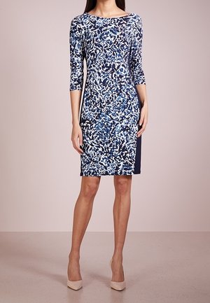 Femme portant une robe ajustée mi-longue à imprimé abstrait bleu, noir et blanc, avec des manches trois-quarts, associée à des escarpins beige.