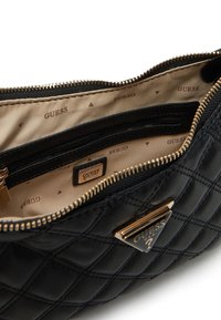 Borsa a tracolla nera trapuntata con hardware dorato, dotata di chiusura a zip. L'interno presenta una fodera beige semplice con un motivo ripetuto del logo "GUESS".