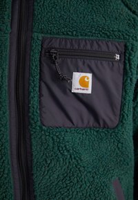 Fleecejacke in dunkelgrün mit einer schwarzen Nylon-Reißverschlusstasche. Merkmale ein quadratisches Stoffetikett mit einem orangefarbenen Logo. Strukturierten, weichen Oberfläche.