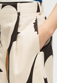 Χέρι στην τσέπη μπεζ παντελονιού με μεγάλα αφηρημένα μαύρα και καφέ σχήματα, ορατή μέση και μέρος ριγέ πουκάμισου από πάνω.