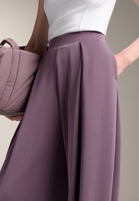 Plissierte lila Hose aus weichem Stoff mit hoher Taille und glatter Verarbeitung. In einer Hand wird eine passende strukturierte Tasche gehalten.
