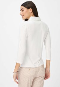 Weißes Langarm-Polohemd mit Kragen, kombiniert mit gemusterten beigen Hosen. Glatte Textur, taillierte Passform und minimalistische Details sichtbar.