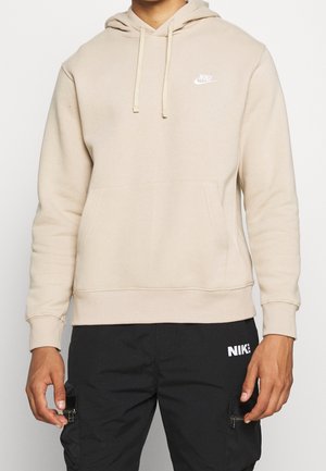 Mężczyzna w beżowej bluzie z kapturem Nike z przednią kieszenią i czarnych spodniach Nike z kieszeniami na suwak po bokach, stojący na białym tle.