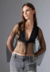 Fekete, rövidített fazonú felső áttetsző, pöttyös anyagból, mély V-nyakkal, hosszú ujjakkal és elöl megkötős részlettel, magas derekú farmernadrág fölött viselve.