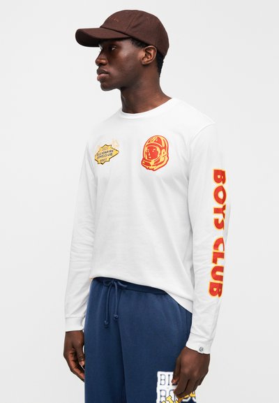 Λευκό μακρυμάνικο μπλουζάκι με κόκκινες γραφιστικές λεπτομέρειες, που απεικονίζει έναν αστροναύτη και το κείμενο «BOYS CLUB» στο μανίκι. Συνδυασμένο με σκούρο μπλε παντελόνι.