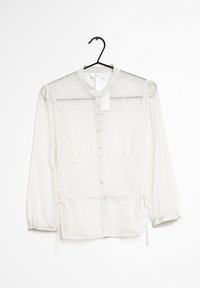 Blusa blanca transparente con cuello redondo, cierre de botones y dobladillo ajustable con cordón. Presenta pequeños lunares negros y mangas largas.