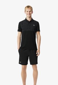 Czarna koszulka polo z wzorzystą górną częścią; krótkie rękawy i dopasowane czarne szorty uzupełniają wygląd. Widoczne logo Lacoste.