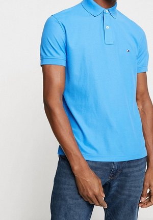Mężczyzna ubrany w jasnoniebieską koszulkę polo z krótkim rękawem i małym logo na piersi oraz ciemnoniebieskie dżinsy.