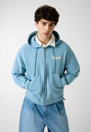 Giovane uomo con felpa azzurra chiara con zip sopra camicia bianca con colletto, mani nelle tasche, abbinata a jeans blu larghi, sfondo neutro.