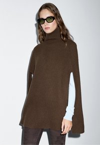 Donna che indossa un ampio mantello marrone a costine con collo alto e spacchi che mostrano le maniche azzurro chiaro, pantaloni scuri e grandi occhiali da sole con lenti verdi.