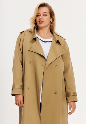 Femme portant un trench-coat beige croisé sur une chemise blanche avec un motif ovale noir près de l'encolure, debout devant un fond clair.