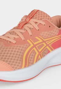 Ανοιχτό κοραλί αθλητικό παπούτσι ASICS με κίτρινες ρίγες λογότυπου, επάνω μέρος από δίχτυ, επενδυμένο γιακά και λευκή σόλα με κοραλλί λεπτομέρειες.