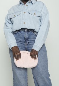 Jasno niebieska, skrócona kurtka dżinsowa z dwoma kieszeniami na piersi, zestawiona z wysokim stanem niebieskich dżinsów. Trzyma miękką, różową, sztuczną futrzaną kopertówkę.