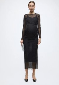 Rochie de maternitate neagră cu mâneci lungi din material transparent, detalii adunate la nivelul burții, tiv până la genunchi și accente strălucitoare subtile pe întreaga suprafață.