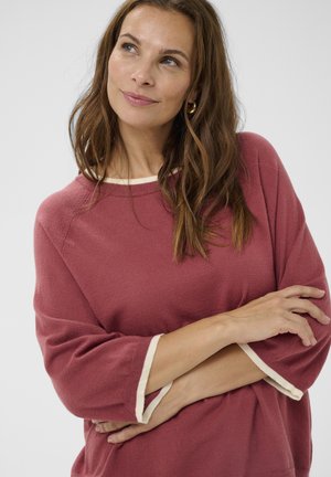 Frau mit langen braunen Haaren, die einen staubigen rosafarbene Pullover mit weißen Bündchen trägt, die Arme verschränkt und nach oben schaut, vor einem schlichten hellen Hintergrund.