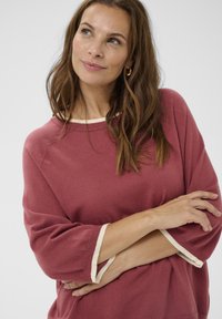 Hosszú barna hajú nő poros rózsaszín pulóvert visel, amelynek fehér mandzsettái vannak, karba tett kézzel, felfelé néz egy egyszínű világos háttér előtt.