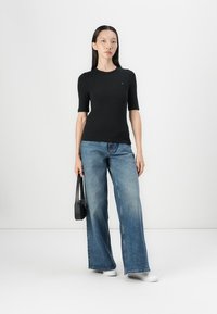 Zwarte korte mouwen top gecombineerd met lichtblauwe wijde jeans. Accessoires zijn een kleine zwarte tas. Model staat voor een effen achtergrond.