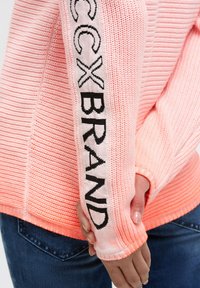 Knit sweater in light pink with textured pattern; features "CCX BRAND" embroidered in black on the sleeve. Ribbed cuffs and hem.