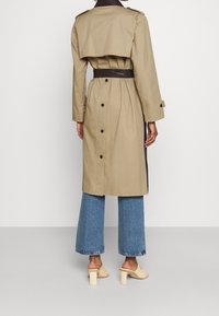 Kobieta w beżowym trenczu z czarnym paskiem, w niebieskich, krótkich dżinsach i kremowych klapach na obcasie, stojąca plecami do białego tła.