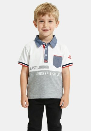 Jeune garçon aux cheveux blonds portant un polo blanc et gris avec le texte "East London" et un pantalon noir, debout et souriant.