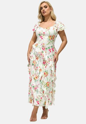 Frau trägt ein weißes Midi-Kleid mit Blumenmuster, Rüschenärmeln und mehrfachen Rüschen, posiert mit den Händen nahe der Hüften vor einem schlichten Hintergrund.