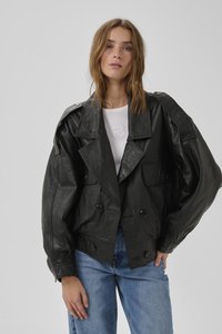 Vrouw die een zwarte oversized leren jas draagt over een wit topje en blauwe jeans, staand tegen een eenvoudige lichtgrijze achtergrond.