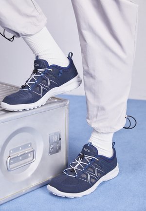 Zapatillas deportivas de color azul marino con paneles de malla, detalles en blanco y cordones. Llevadas con calcetines blancos, descansando sobre una maleta plateada.