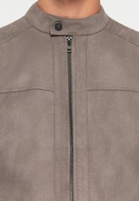 Chaqueta de ante gris con un alto cuello, cierre central de cremallera y detalles de costura sutiles en los hombros y el cuerpo.