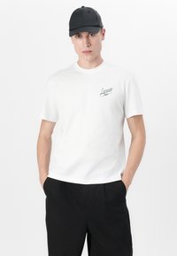 Balta medvilnės marškinėliai su žalia "Lacoste Paris" logotipu kairėje krūtinės pusėje, derinami su juodomis kelnėmis ir juodu kepurėliu.