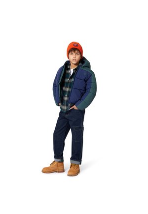 Veste matelassée bleu marine et verte, chemise à carreaux, jean foncé et bottes beiges. Bonnet en tricot orange, coupe décontractée et design structuré.