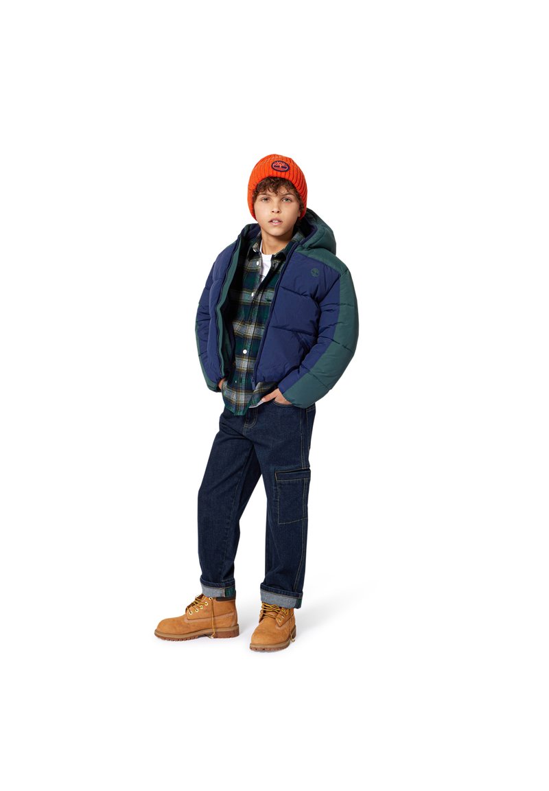 Veste matelassée bleu marine et verte, chemise à carreaux, jean foncé et bottes beiges. Bonnet en tricot orange, coupe décontractée et design structuré.