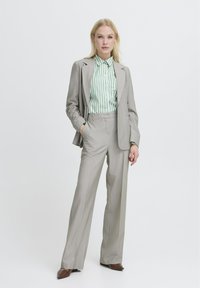 Traje gris claro a medida con pantalones de pierna ancha y una chaqueta a juego, combinado con una camisa de botones a rayas verdes y blancas. Zapatos marrones.