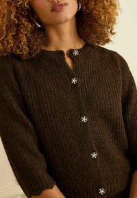 Hnedý pletený sveter so štruktúrou rebrovania, s výstrihom do V a ozdobnými gombíkmi v tvare kvetov zdobenými drahokamami.