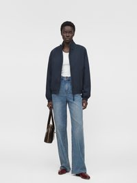 Femme portant une veste bleu marine, un haut blanc, un jean bleu à jambes larges, des chaussures rouges, tenant un sac à main marron, debout devant un fond clair uni.