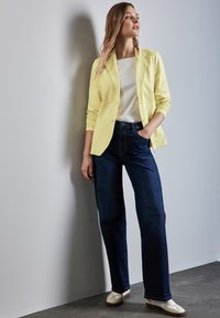Gul skräddarsydd kavaj med långa ärmar över en vit topp, i kombination med mörkblå vida jeans och ljusa skor.