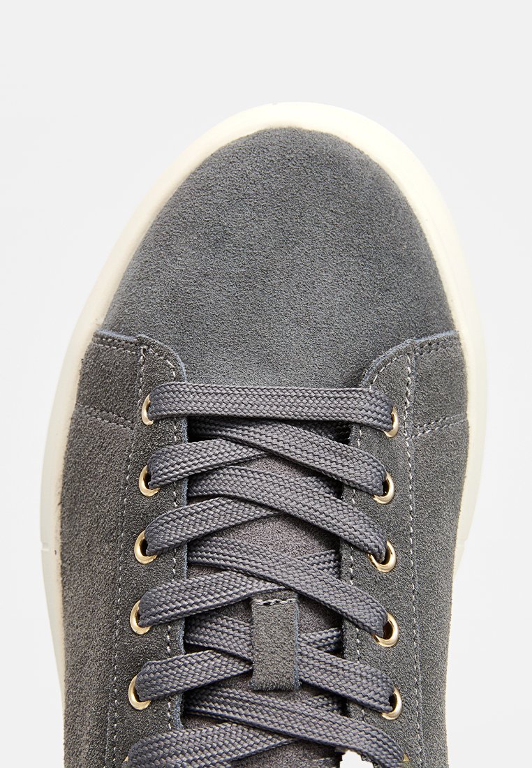 Sneaker in suede grigio con punta rotonda, occhielli dorati e lacci grigi piatti. Presenta una tomaia testurizzata e una suola in gomma bianca a contrasto.