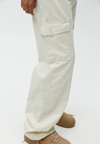 Pantalon cargo beige à coupe ample, texture en tissu, poche latérale avec fermeture par bouton, et détails cousus, associé à des baskets marron.