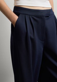 Main dans la poche, portant un pantalon tailleur bleu marine à taille haute plissé et un top blanc à col ribbé, sur fond uni.