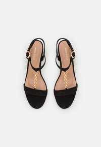 Mustad nahast sandaalid, millel on lai rihm, ankle strap ja kuldne ümmargune klamber, samuti dekoratiivne kuldne ketiasend varvaste kohal.
