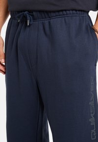 Navyblauwe sweatpants van zacht materiaal, met een elastische tailleband en een trekkoord, en een zijlogo in lichtgrijs.