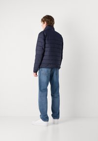 Jūros spalvos pūkinė striukė su horizontaliais siuvinėtais raštais, aukštu apykakle, derinama su šviesiai mėlynomis džinsinėmis kelnėmis ir baltomis sportinėmis bateliais, matoma iš nugaros.