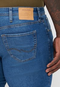 Denimjeans i mellomblå med et lærmerke merket "JACK & JONES." Har en rund baklomme med subtile stikningdetaljer.