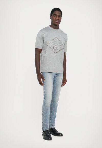 Pilka trumpa rankovė t-shirt su deimanto logotipu ir sparnais, viduryje esantis "GA", derinama su šviesiai mėlynomis džinsomis ir juodais batais.
