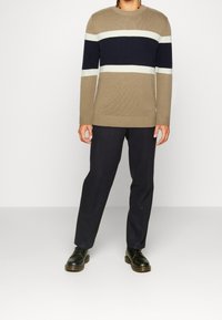 Beżowy sweter z prążkowanej dzianiny z granatowo-białymi poziomymi paskami, w połączeniu z ciemnymi spodniami i czarnymi sznurowanymi butami.