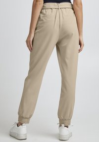 Beige Joggerhosen mit elastischen Bündchen, aus einem glatten, leichten Stoff und in einer lockeren Passform, getragen mit weißen Sneakers.