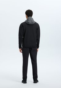 Giacca nera con cappuccio grigio, texture liscia e tasche con zip. Abbinata a pantaloni neri e scarpe scure, vista da dietro.