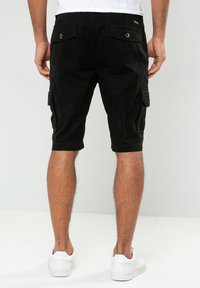 Pantalones cortos de carga negros con bolsillos de parche, hechos de algodón, de longitud hasta la rodilla, con detalles de botones y una tela texturizada. Llevados con zapatillas blancas.