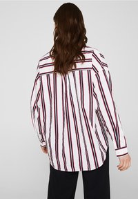 Camicia bianca a maniche lunghe con strisce verticali rosse e blu navy, caratterizzata da un dettaglio a bottone ai polsini e una vestibilità comoda.